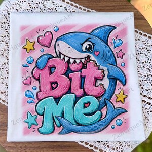May include: White t-shirt featuring a cartoon shark biting the words "Bit Me". The shark is blue with white teeth and a pink heart on its cheek. Pink and blue accents include hearts, stars, and water droplets.