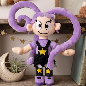 Könnte beinhalten: Eine Plüschpuppe mit hellvioletter Haut und langem, gelocktem violettem Haar. Die Puppe trägt ein violettes Oberteil unter einer schwarzen Latzhose mit einem gelben Stern auf der Brust und gelben Sternen auf der schwarzen Leggings sowie violette Schuhe.