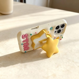 May include: A cream-colored phone case with a yellow star-shaped phone grip. The case has the word "STAR" in pink letters and is decorated with colorful confetti. The phone case is designed to fit a smartphone.