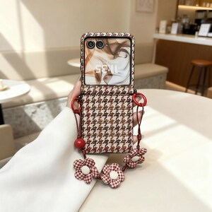Op de afbeelding: Een telefoonhoesje met een bruin en crème pied-de-poule patroon, met een bandje versierd met gingham bloemen. Het telefoonscherm toont de tijd 11:35. De band en de hoes hebben rode accenten.