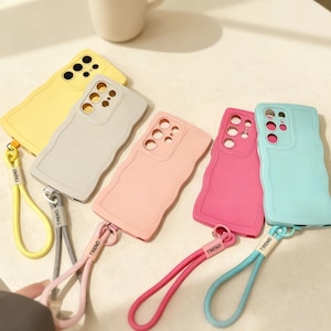 Puede incluir: Una colección de fundas de teléfono coloridas en amarillo, gris, rosa, fucsia y azul. Cada funda tiene un diseño de borde ondulado y una correa de muñeca a juego con la palabra "TREND" impresa. Las fundas están diseñadas para un modelo de teléfono específico.