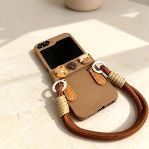 Può includere: Una custodia per telefono marrone con cinturino, decorata con ciondoli a forma di biscotto e chicchi di caffè. La custodia ha un design pieghevole e un cinturino marrone abbinato con dettagli beige. La custodia è progettata per un telefono pieghevole.