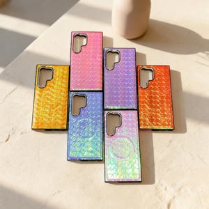 May include: A collection of colorful phone cases in shades of yellow, pink, purple, blue, and orange. Each case has a textured, iridescent finish and a black border. The cases are arranged on a light-colored surface.