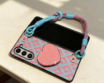 May include: A phone case with a pink heart-shaped grip and a blue and pink patterned design. The case has a matching blue and pink strap. The case has a camera with three lenses.
