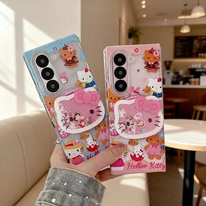 May include: Two phone cases featuring Hello Kitty designs. One case is light blue, the other is pink. Both cases have a pop-up Hello Kitty face and are decorated with various Hello Kitty characters and accessories. The text "Hello Kitty" is visible on the pink case.