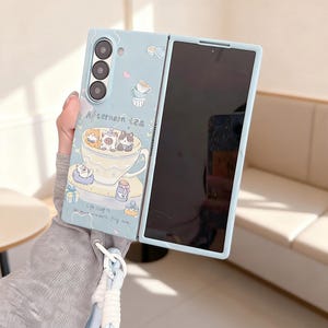 Könnte beinhalten: Hellblaue Handyhülle mit einem skurrilen Design. Sie zeigt eine Teetasse mit Cartoon-Tieren und den Worten "Afternoon Tea". Die Hülle ist geöffnet und zeigt einen schwarzen Bildschirm. Das Design umfasst Herzen, Geschenke und andere Tee-Elemente.