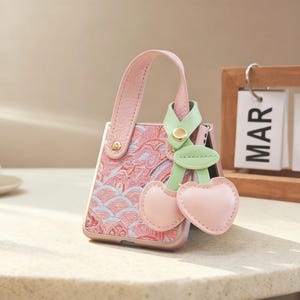 Puede incluir: Funda de teléfono rosa con asa a juego y charm de cereza. La funda presenta un patrón de ondas rosas y azules. El charm es de color verde claro y rosa. Un calendario de madera muestra el mes de marzo.