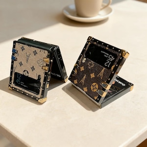 May include: Two open, black and gold phone cases with a brown and tan pattern. The cases are on a light-colored surface. The phone screens display the time. The cases have gold corner accents and clasps.