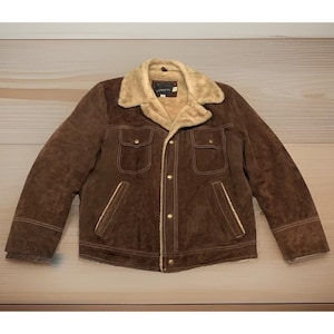 Puede incluir: Una chaqueta de ante marrón con cuello y forro interior de piel sintética beige. La chaqueta incluye dos bolsillos en el pecho con solapas abotonadas, dos bolsillos laterales y un cierre frontal con botones metálicos y costuras claras visibles.