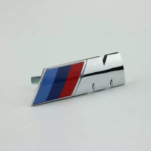 Pode incluir: Um emblema M metálico com um design inclinado e três faixas diagonais distintas em azul claro, azul escuro e vermelho. O emblema tem um acabamento cromado polido e uma forma angular nítida, representando um logótipo automóvel reconhecível.