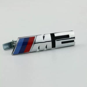 Pode incluir: Um emblema de metal com acabamento cromado apresentando o texto M2 em uma fonte de bloco em negrito. À esquerda das letras, há uma seção retangular inclinada com três listras diagonais em azul claro, azul escuro e vermelho. Um clipe está fixado.