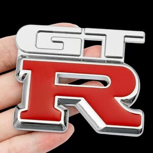 Puede incluir: Un emblema GT-R cromado y rojo. Las letras "GT" son cromadas y la letra "R" es roja. El emblema tiene un acabado brillante y probablemente sea para un coche.