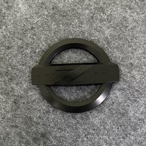 Könnte beinhalten: Ein schwarzes Autoemblem mit einem kreisförmigen Design und einem rechteckigen Balken in der Mitte. Das Emblem hat eine glänzende Oberfläche und ist wahrscheinlich aus Metall. Der Hintergrund ist eine graue, strukturierte Oberfläche.