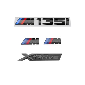 Pode incluir: Um conjunto de emblemas automotivos composto por um emblema M135i preto com um logótipo M de listras azuis, roxas e vermelhas, dois logótipos M menores com o mesmo esquema de cores e um emblema XDrive preto com acabamento brilhante e letras em relevo.