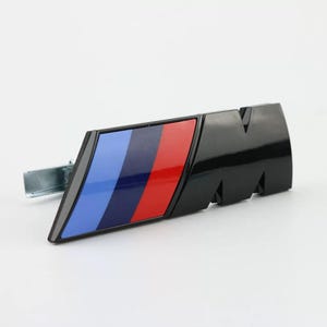 May include: A glossy black M emblem badge featuring a diagonal stripe pattern in light blue, dark blue, and red. The automotive accessory has a metallic mounting bracket attached to the left side, designed for exterior vehicle attachment.
