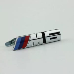 Pode incluir: Um emblema M6 com acabamento cromado apresentando um design inclinado com três listras diagonais distintas em azul claro, azul escuro e vermelho no lado esquerdo. As letras metálicas M e 6 são exibidas em destaque em estilo de bloco.