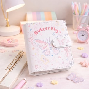 Puede incluir: Diario blanco con diseño de mariposa. La portada presenta una gran ilustración de mariposa rosa y azul y la palabra "Butterfly". Un pequeño despertador rosa y bolígrafos están en el fondo.