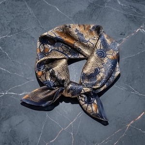 May include: A square silk scarf featuring a repeating pattern of dark blue and metallic gold abstract shapes and wave-like lines. The fabric has a smooth, reflective sheen and is tied into a loose knot, displaying intricate geometric details.