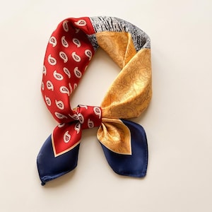 May include: A square silk scarf tied in a knot, featuring a multi-colored patchwork design. The pattern includes sections of red with white paisley, yellow with intricate gold floral motifs, black and white geometric lines, and solid navy blue.