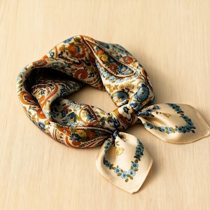 May include: A square silk scarf featuring a cream backdrop and a paisley floral design in orange, blue, and brown hues. The scarf is knotted, with two cream-coloured ends decorated with blue floral borders.