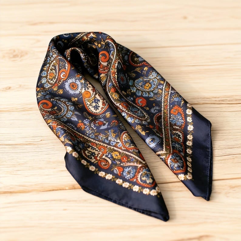 May include: A navy blue silk scarf featuring a paisley design in orange, blue, and brown hues. The square scarf has a dark blue border. It is folded on a wooden surface.