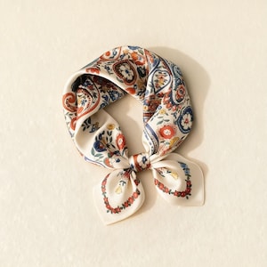 Pode incluir: Um lenço de seda creme com um padrão paisley e floral em vermelho, azul e amarelo. O lenço é amarrado em um nó, com as pontas formando um laço. O lenço tem formato quadrado.