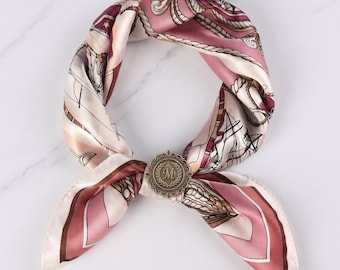 May include: A square silk scarf in shades of dusty pink, cream, and dark brown, featuring a nautical print with ropes and sailing ships. The scarf is secured with a round, antique brass-toned metal clip embossed with the letter M.