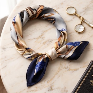 May include: A square silk scarf tied in a loop, featuring a pattern of gold chains, buckles, and blue floral motifs on a light gray and cream background, with solid dark navy blue corners and edges.