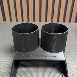 Könnte beinhalten: Ein schwarzer Doppel-Getränkehalter mit zwei zylindrischen Öffnungen und einer Basis mit dem geprägten Text CRX Si. Das Objekt hat eine matte Oberfläche und ein strukturiertes Design mit zwei abgewinkelten Stützbeinen an der Vorderseite.