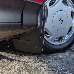 Pode incluir: Um close-up do lado de um carro vermelho, com um guarda-lamas cinzento com a palavra "HONDA" em relevo. O carro tem uma roda prateada com o logótipo da Honda e um pneu preto. O carro está estacionado numa superfície cinzenta texturizada.