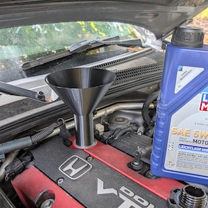 Op de afbeelding: Een zwarte plastic trechter in de olievulopening van een rode Honda DOHC VTEC kleppendeksel, naast een blauwe fles van 1 liter SAE 5W-40 volledig synthetische motorolie met een zwarte dop die op het motoroppervlak rust.