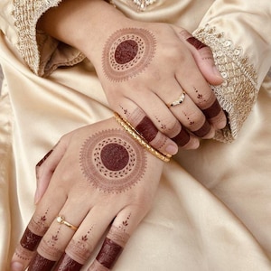 May include: Hands adorned with intricate henna designs. The palms feature large, circular patterns with a dark reddish-brown center, surrounded by detailed, lighter brown motifs. Fingers are decorated with matching henna bands and delicate dot patterns, complemented by gold rings and a gold bracelet.