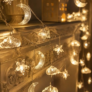 May include: A string of LED fairy lights featuring clear, textured plastic shapes of crescent moons, stars, and clouds. Each individual ornament is illuminated from within, emitting a warm, soft glow along the thin, flexible wire cord.