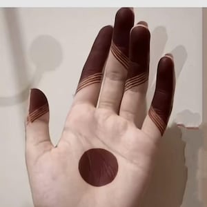 May include: A hand with henna designs. The fingers and palm are decorated with a dark reddish-brown henna. The fingers have striped patterns, and the palm features a large, solid circle. The background is a plain, light color.