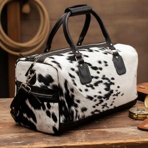 May include: A duffel bag with a cowhide pattern in black and white. The bag has black leather handles, trim, and a shoulder strap. The bag is rectangular and appears to be made of a durable material, suitable for travel or carrying luggage.