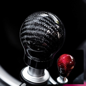 Puede incluir: Un pomo de cambio de fibra de carbono negro con un acabado brillante. El pomo es redondo y tiene un patrón tejido. También se ve un pomo de cambio de fibra de carbono rojo más pequeño.