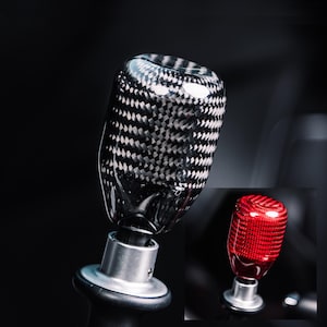 Op de afbeelding: Een versnellingspookknop van koolstofvezel met een zilveren basis. De knop heeft een geweven zwart-wit patroon. Een kleinere, rode versnellingspookknop is rechtsonder te zien. Auto-accessoire.