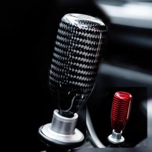 May include: A close-up of a black and gray carbon fiber shift knob with a silver base. The shift knob has a glossy finish and a woven pattern. A smaller red shift knob is visible in the background.