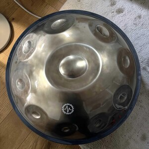 May include: A silver-colored steel handpan drum with a dark blue rubber rim. The instrument has multiple circular indentations on its surface, with a central indentation and a logo near the bottom. The handpan is designed for melodic percussion.