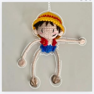 Op de afbeelding: Gehaakte Luffy-pop met een gele strohoed, rood vest en blauwe korte broek. De pop heeft een lachend gezicht en lange, bungelende armen en benen. Opgehangen aan een ketting.