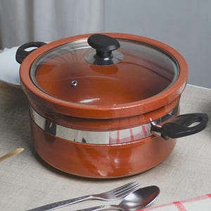May include: A terracotta-coloured cooking pot with a glass lid and black handle. The pot has a stainless steel band around the middle. A fork and spoon are placed near the pot on a light-coloured surface.