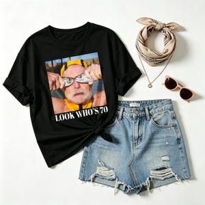 May include: A black t-shirt featuring a graphic of a person wearing a yellow swim cap and holding two fish over their eyes, with the text LOOK WHO'S 70 printed below. Also included are blue denim distressed shorts, a beige patterned bandana, and sunglasses.