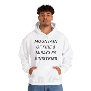 以下が含まれることがあります： 黒い文字で「MOUNTAIN OF FIRE & MIRACLES MINISTRIES」と書かれた白いフード付きスウェットシャツ。スウェットシャツにはフロントポケットとドローストリングフードが付いています。モデルはブルージーンズを着用しています。