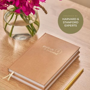 May include: A tan hardcover journal with the text gratitude & potential embossed on the cover, accompanied by a gold pen. A circular green badge reads ENDORSED BY HARVARD & STANFORD EXPERTS. A glass vase with bright pink flowers sits nearby.