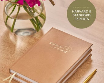 May include: A tan hardcover journal with the text gratitude & potential embossed on the cover, accompanied by a gold pen. A circular green badge reads ENDORSED BY HARVARD & STANFORD EXPERTS. A glass vase with bright pink flowers sits nearby.