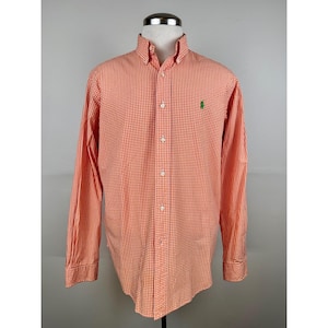 Könnte beinhalten: Orange-weiß kariertes Button-Down-Hemd mit langen Ärmeln. Das Hemd hat einen Button-Down-Kragen und ein grünes Logo auf der Brust. Das Hemd wird auf einer Schaufensterpuppe präsentiert.