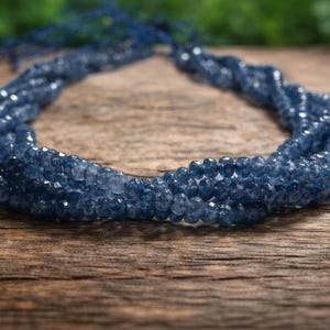Op de afbeelding: Een ketting met meerdere strengen, samengesteld uit kleine, gefacetteerde blauwe kralen. De ketting is gerangschikt in een gedraaid ontwerp en rust op een rustiek houten oppervlak. De achtergrond toont wazig groen gebladerte.