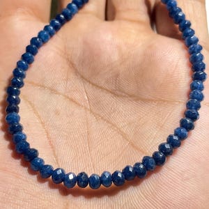 Könnte beinhalten: Eine Kette aus kleinen, facettierten, tiefblauen Edelsteinperlen, die kreisförmig angeordnet sind. Jede einzelne Perle weist eine facettierte Oberfläche auf, die das Licht reflektiert und eine gleichmäßige Textur entlang der gesamten Kette erzeugt.