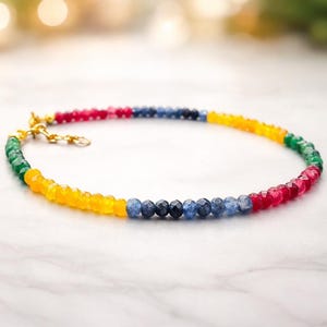 Op de afbeelding: Een veelkleurige kralenketting of armband met kleine, gefacetteerde, ronde edelstenen in de kleuren rood, blauw, geel en groen. Het sieraad is voorzien van een goudkleurige metalen sluiting en een korte verlengketting voor verstelbare lengte.