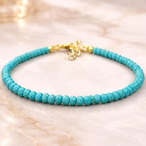 Könnte beinhalten: Ein zartes Armband mit kleinen, facettierten Türkisperlen. Die Perlen haben eine leuchtende türkisfarbene Farbe, und der Verschluss und die Verbindungsglieder sind goldfarben. Das Armband wird auf einer hellen, marmorähnlichen Oberfläche präsentiert.